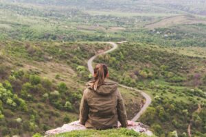 Young person on a high place in nature, looking at a winding road below whose end we don't see.