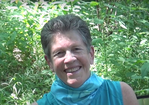 a white woman with short gray in a teal shirt and scarf hair smiles. There are several leaves in the background