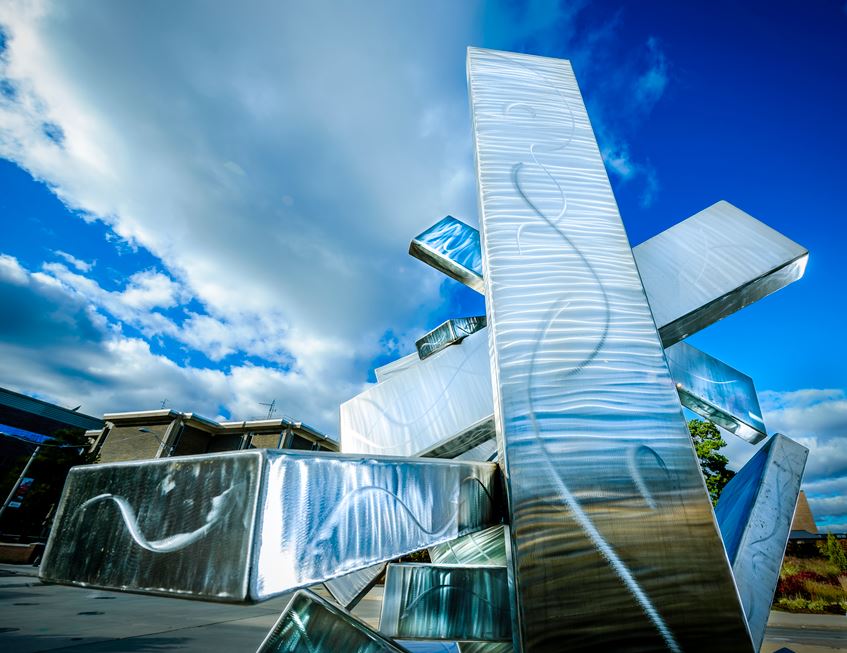 a mirror sculpture sits outside amongst clouds, the reflection of the sky is seen on the sculpture